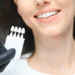 veneers philadelphia pa Close-up of a smiling woman at a dental clinic undergoing a veneer shade selection process. A dentist wearing black gloves holds a shade guide to match the perfect veneer color for a bright, natural-looking smile.