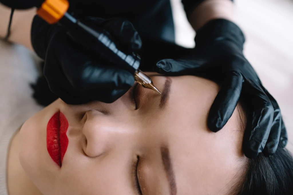 A professional cosmetologist applying permanent eyebrow makeup to a woman's closed eyelids using a micropigmentation tool, highlighting the precision and benefits of permanent makeup in Philadelphia
