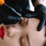 A professional cosmetologist applying permanent eyebrow makeup to a woman's closed eyelids using a micropigmentation tool, highlighting the precision and benefits of permanent makeup in Philadelphia