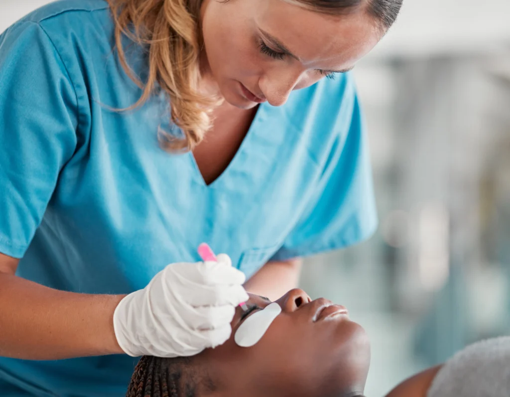 A professional technician in blue scrubs applying eyelash extensions to a client using precision tools, with eye pads in place for protection during the beauty procedure.