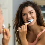 Woman brushing her teeth in front of bathroom mirror smiling